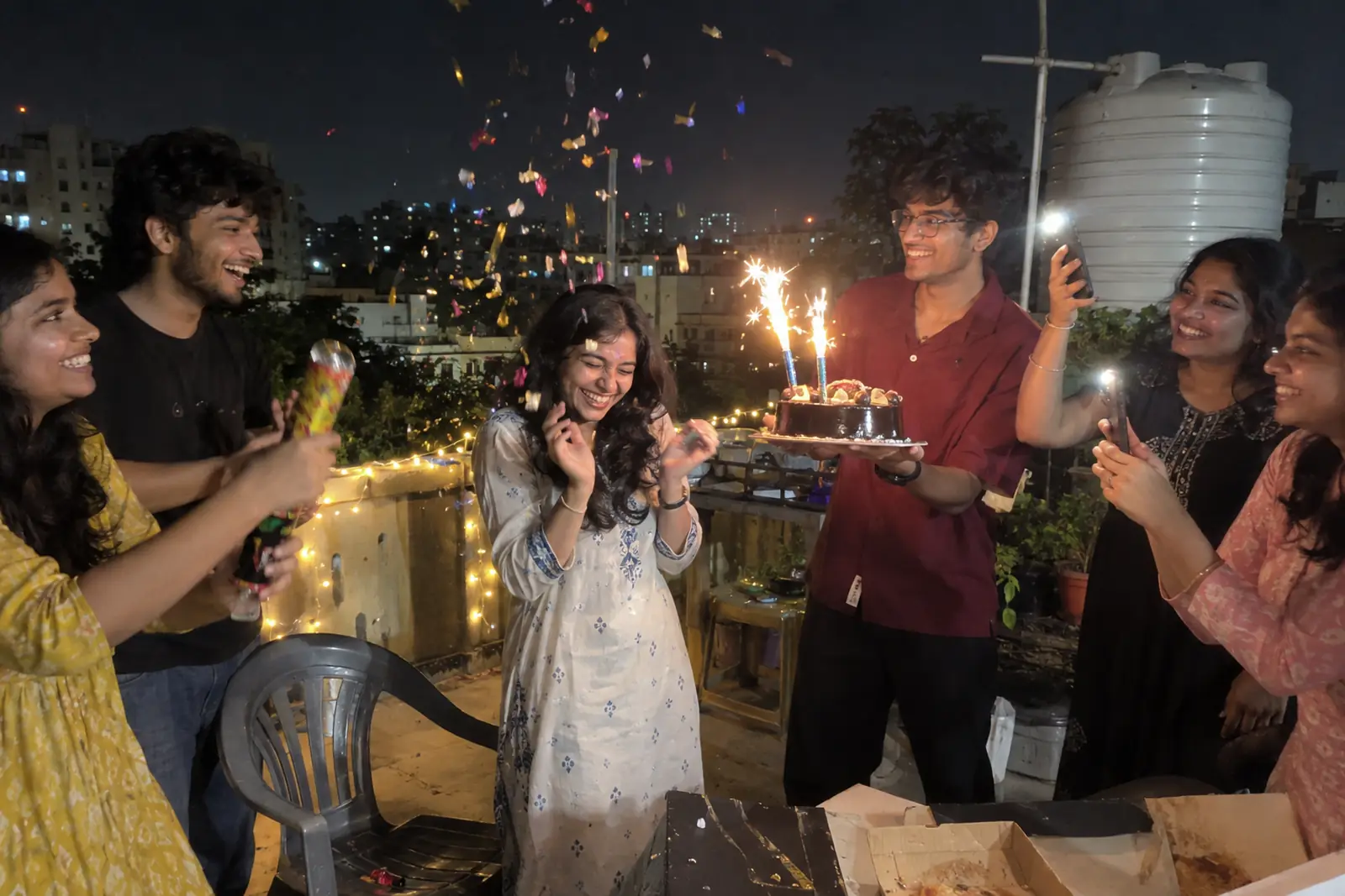 Friends surprise a young woman on a rooftop at night with a sparkler cake and a burst of party poppers