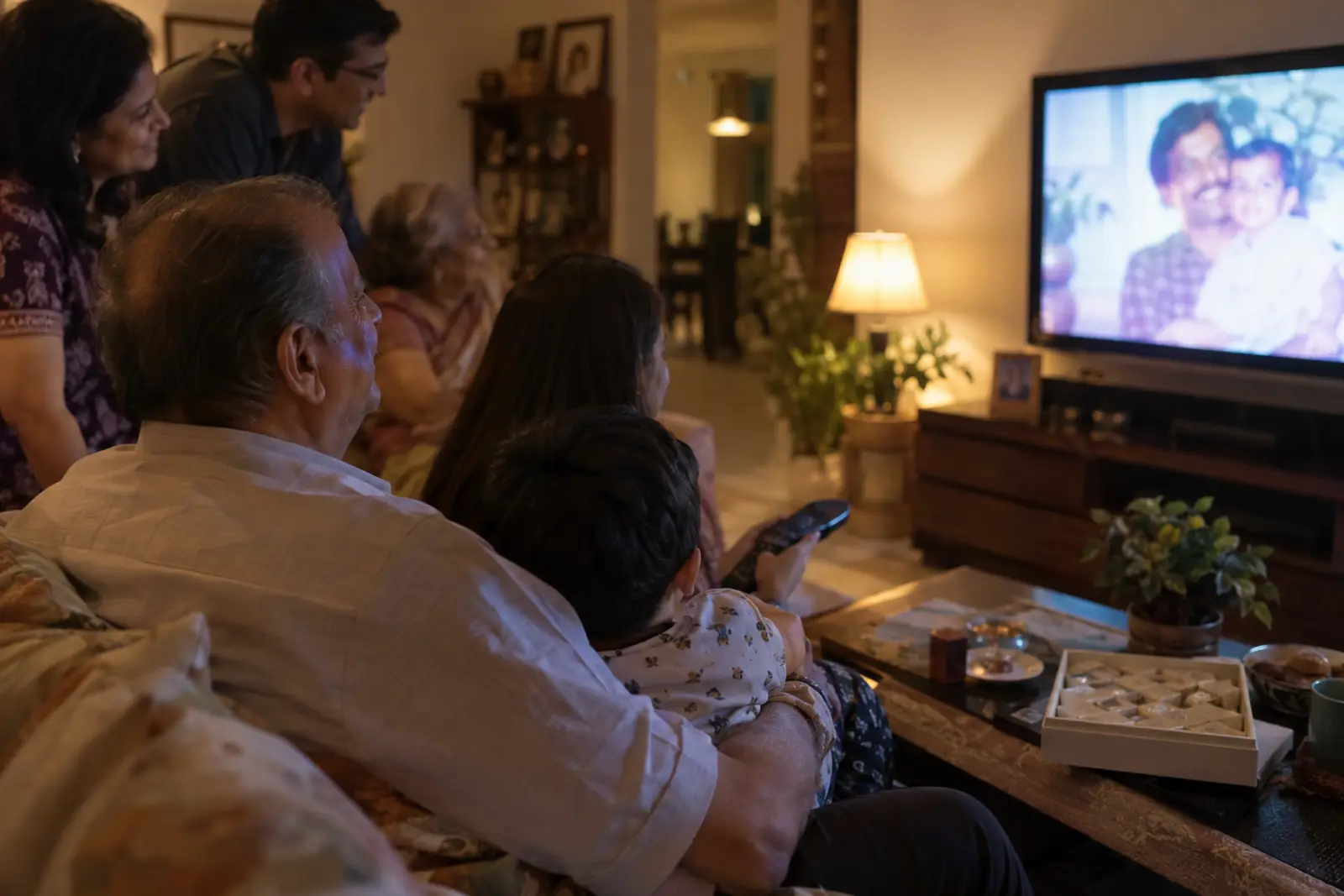 A father turning 60 smiles at old family photos playing on the TV, his grandchild on his lap