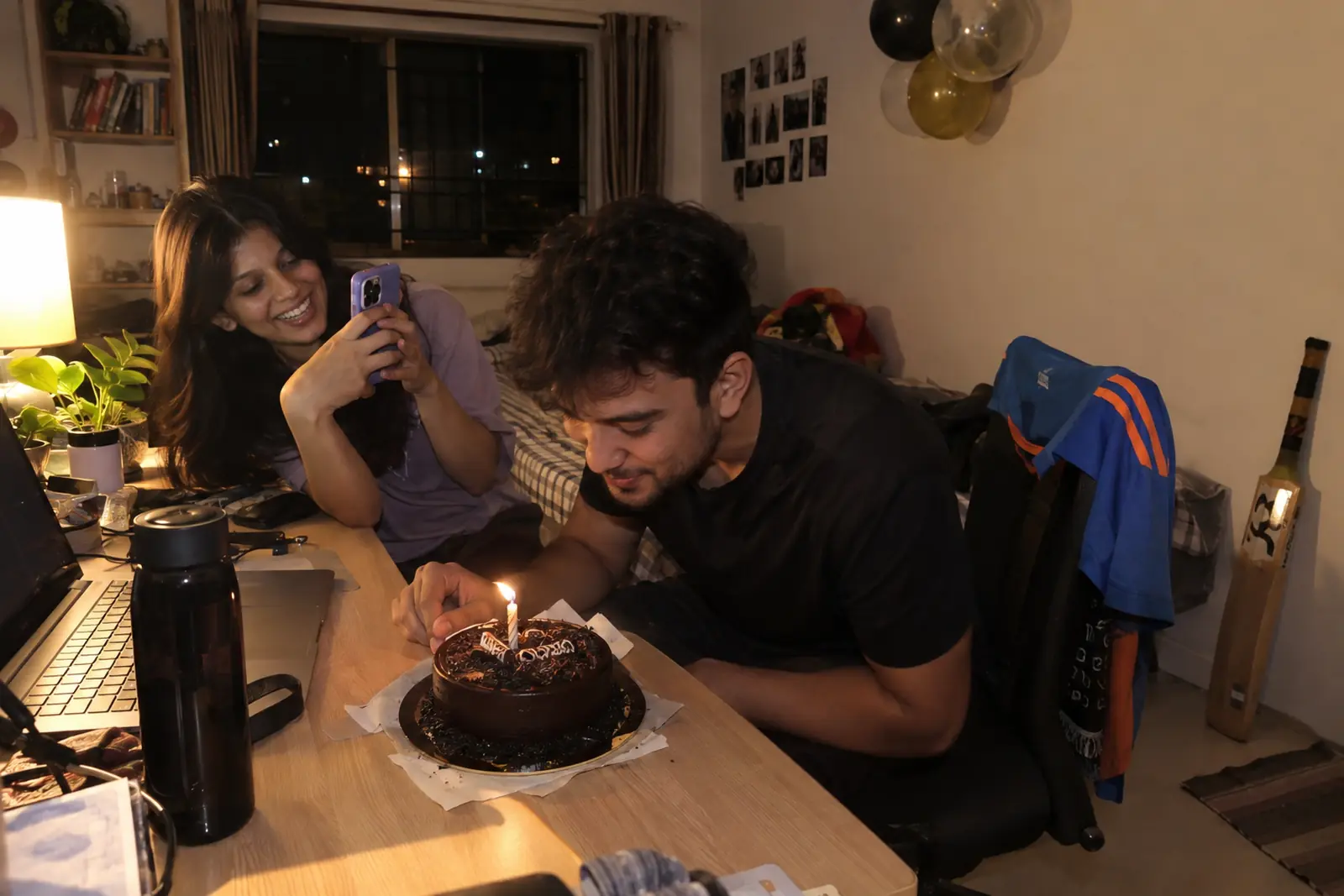 A young man blows out a candle on a chocolate cake at his desk while his girlfriend films him on her phone