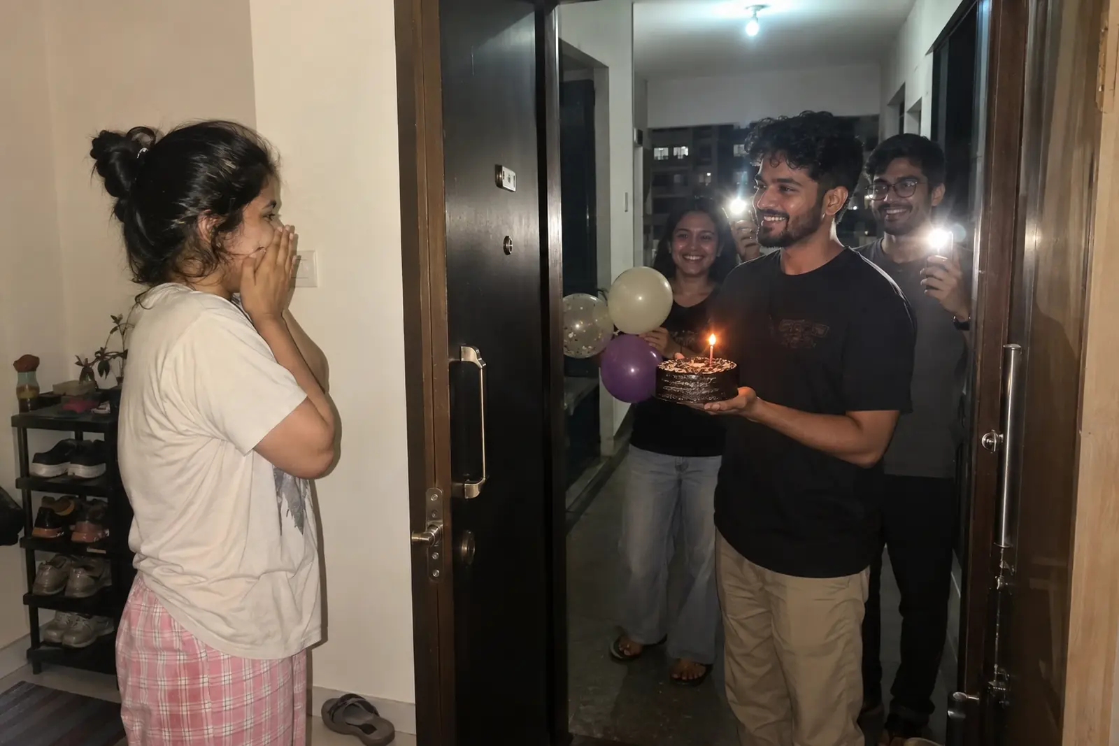 A young woman in pyjamas opens her door at midnight to her boyfriend holding a cake while friends hold balloons and phone torches