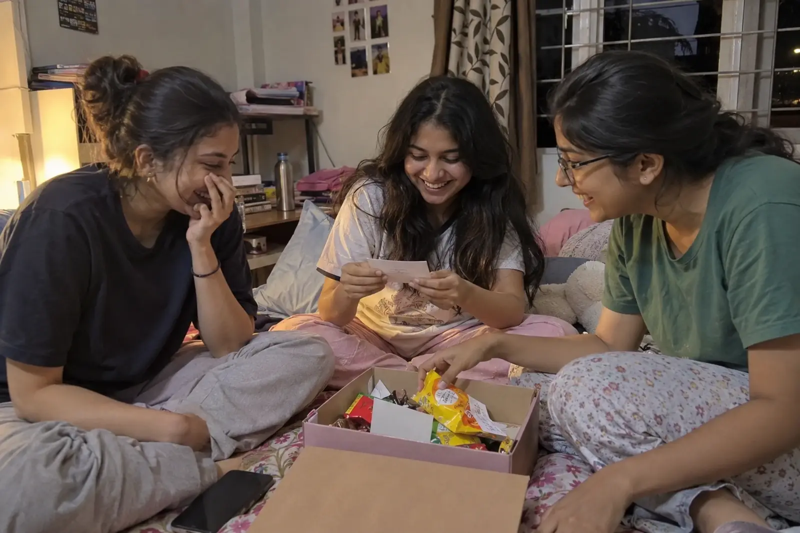 Three college friends on a hostel bed laugh as they open a shoebox of snacks and paper chits
