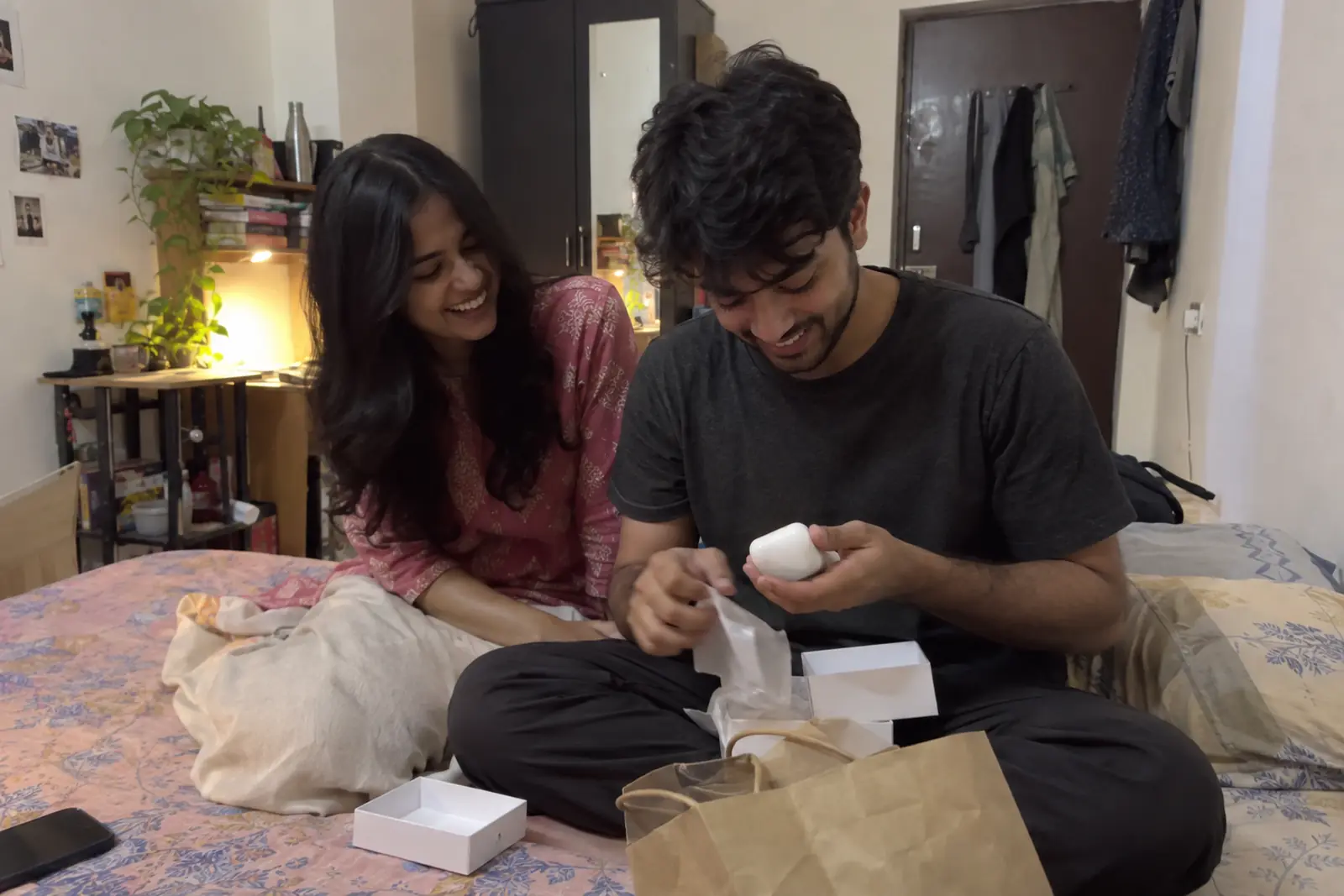 A young man unwraps wireless earbuds on his bed in a rented room while his girlfriend laughs beside him