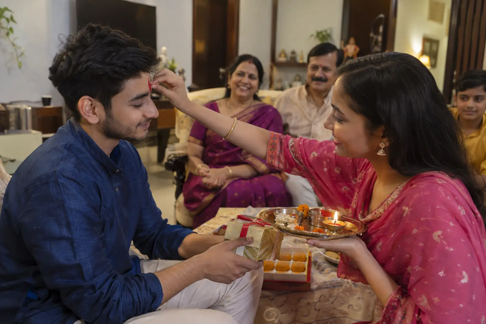 A sister applies a tilak to her brother on Bhai Dooj as he holds a small gift box