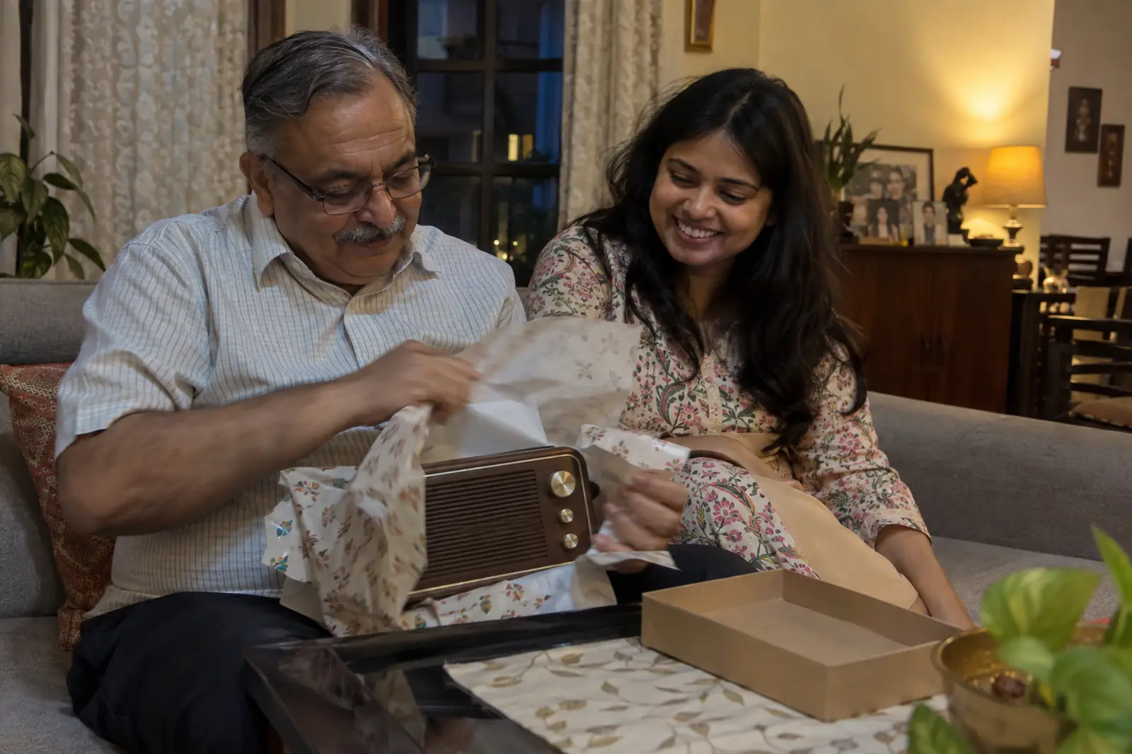 A father unwraps a small retro-style radio speaker on the sofa as his daughter smiles