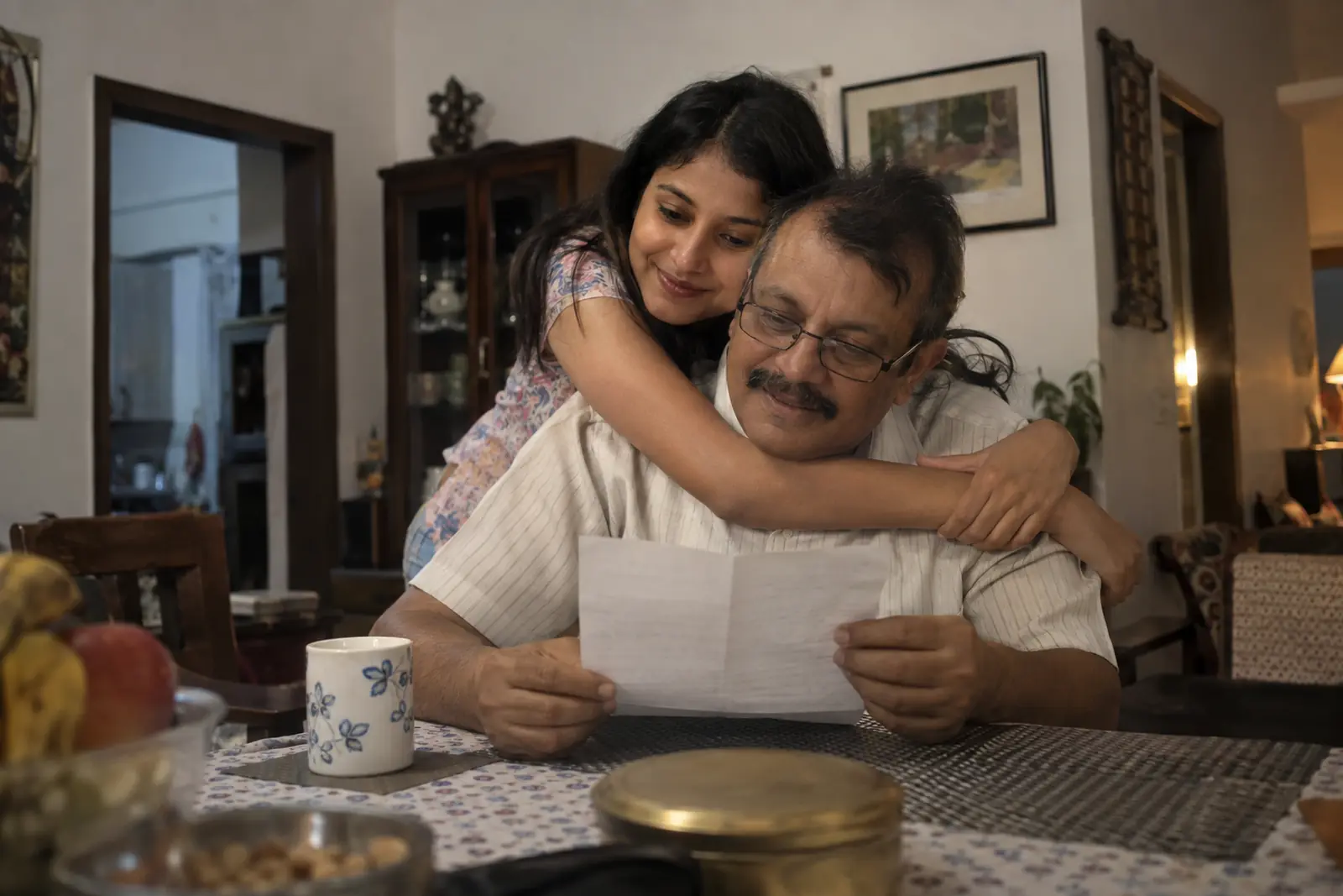A daughter hugs her father from behind as he reads a handwritten letter at the dining table