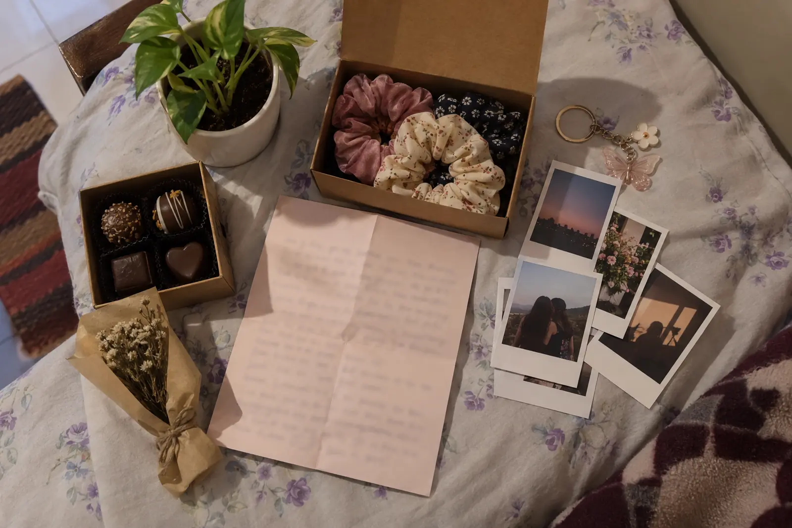 Flat-lay of small gifts for a girlfriend on a hostel bed: scrunchies, instant photos, a letter, chocolates and a tiny plant