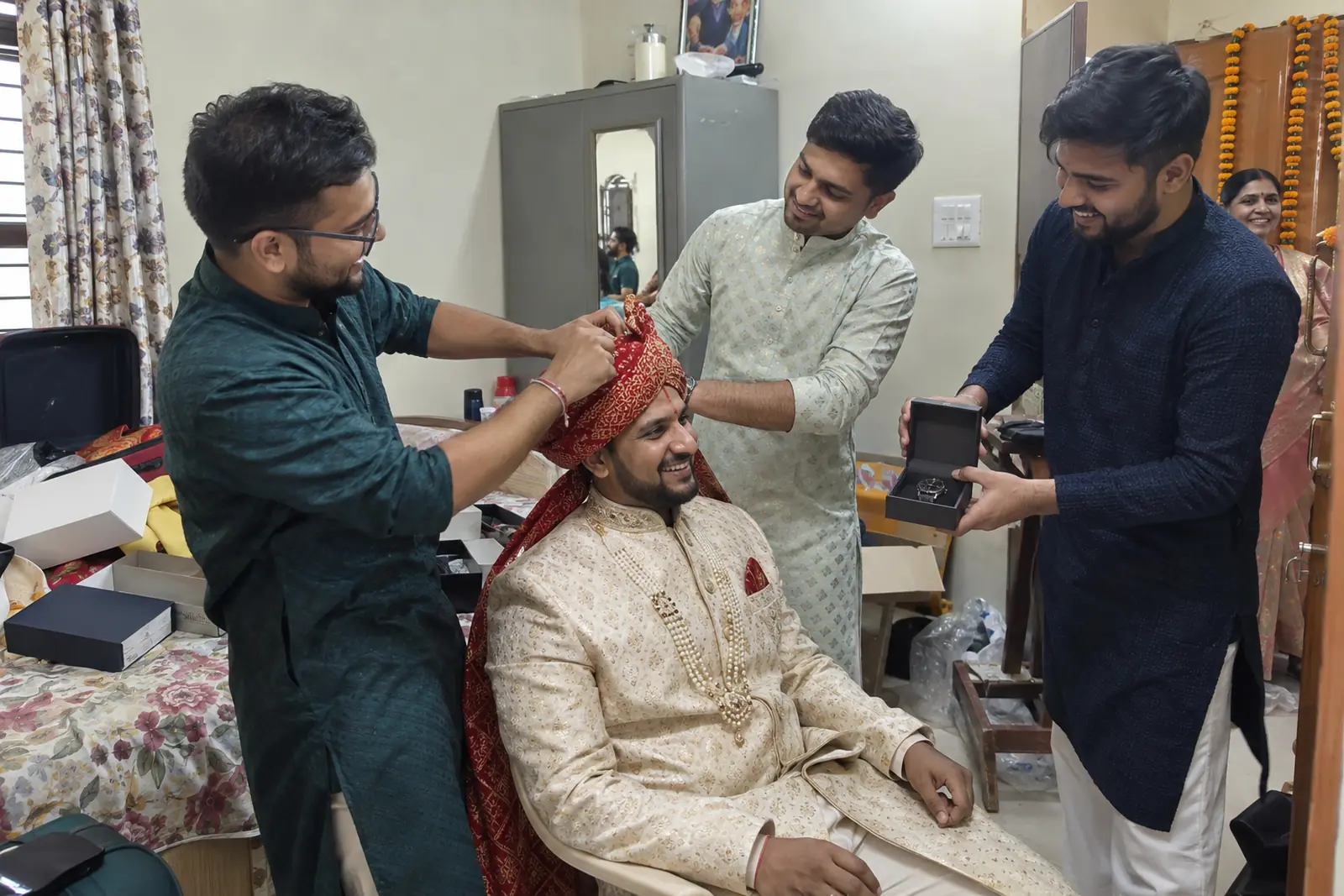 Friends tie a red safa on a groom in a cream sherwani as one hands him an open watch box