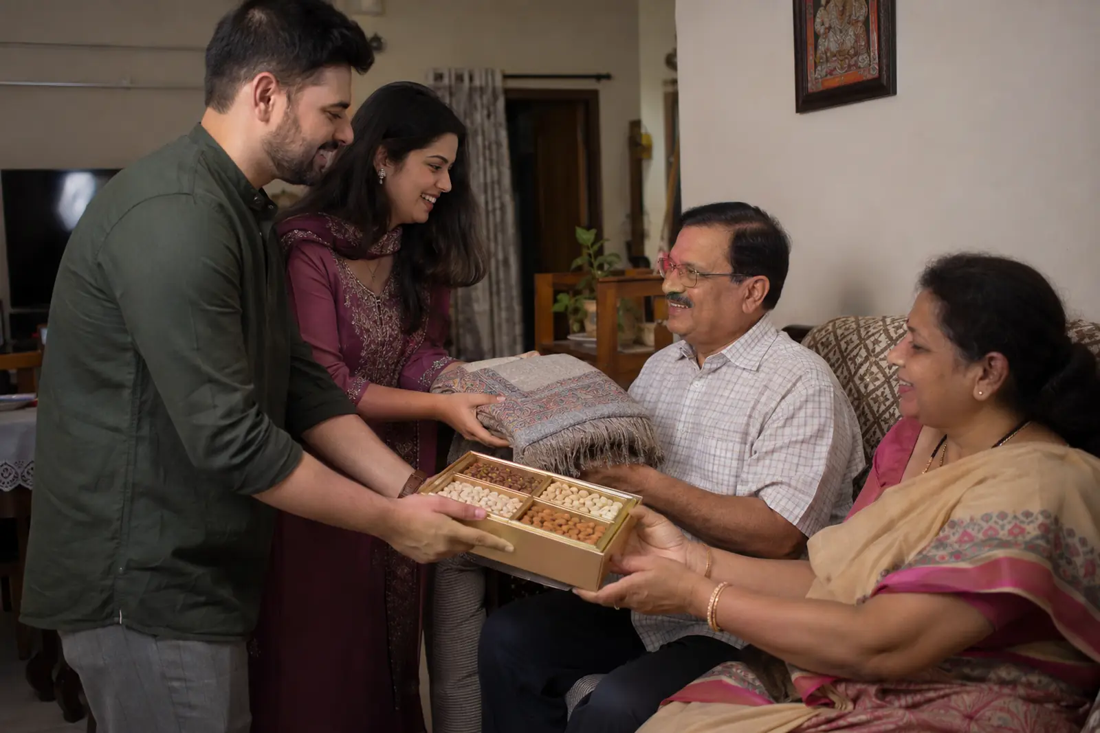 A young couple presents a dry fruit box and a folded shawl to the husband's parents in their living room
