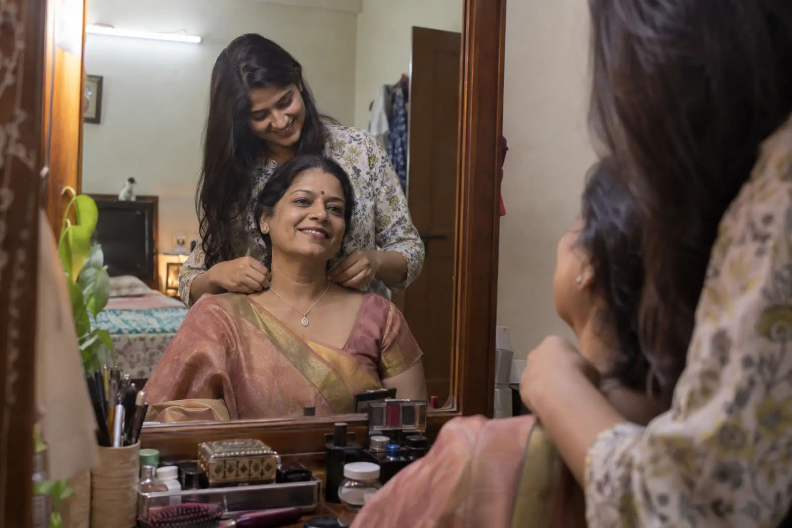 A daughter fastens a silver pendant around her mother's neck in front of a mirror