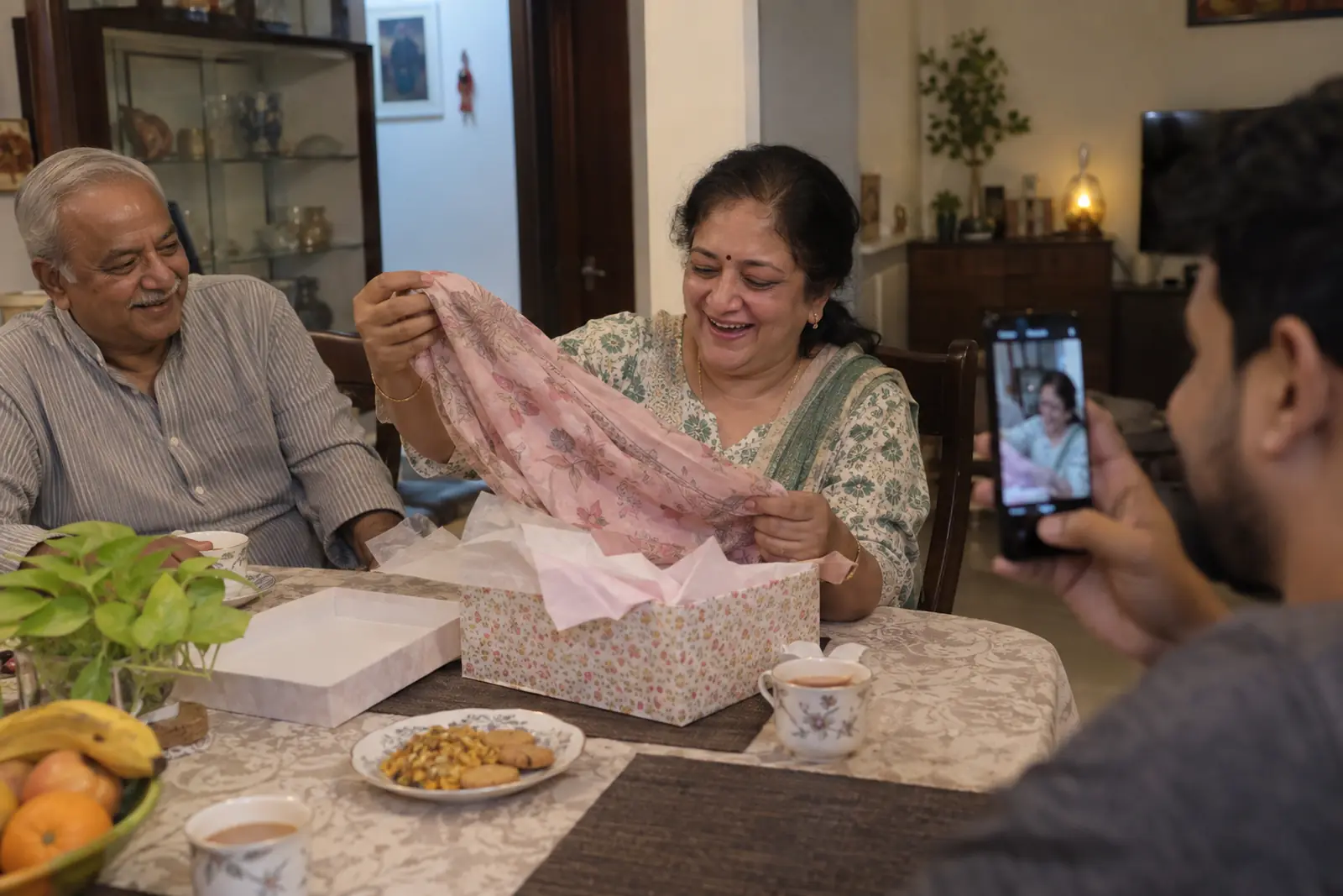 A mother laughs as she unwraps a soft cotton dupatta at the dining table while her son films