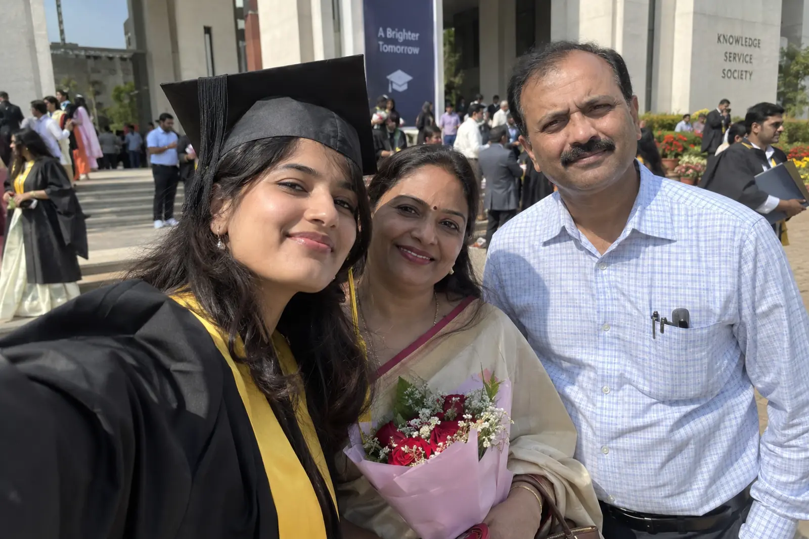 A graduate in a convocation gown and stole takes a selfie with her parents outside her college