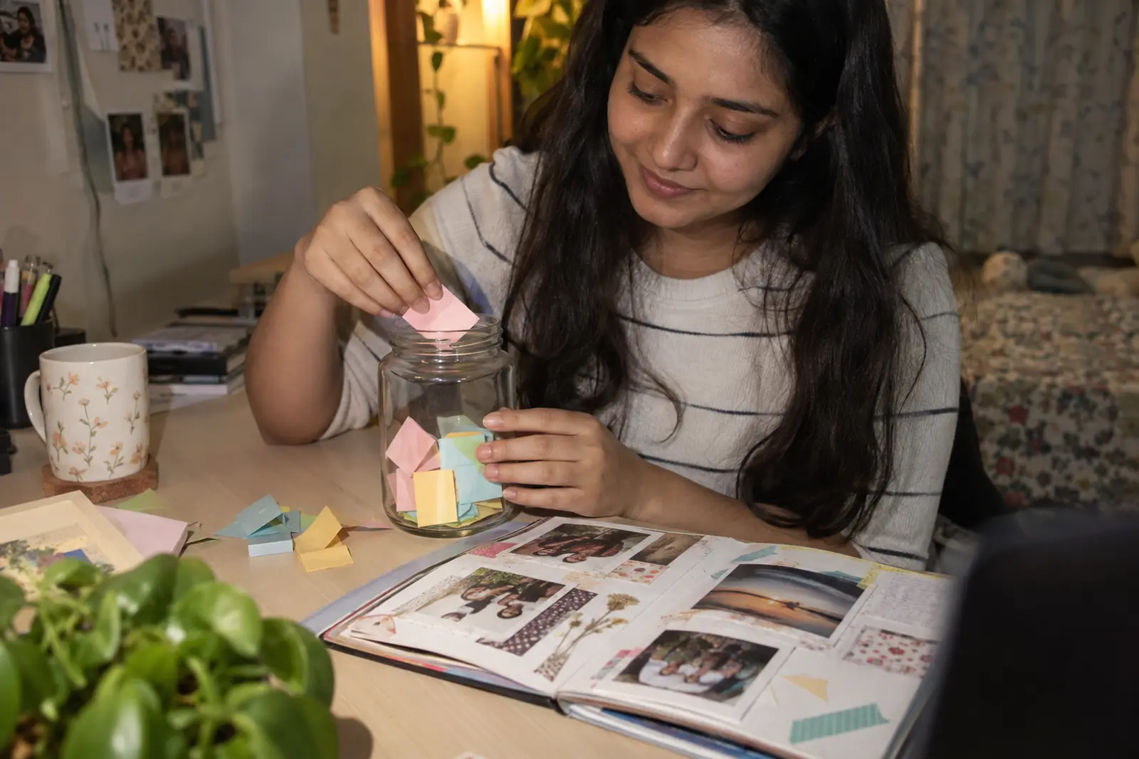 A young woman fills a glass jar with folded notes beside an open scrapbook on her desk