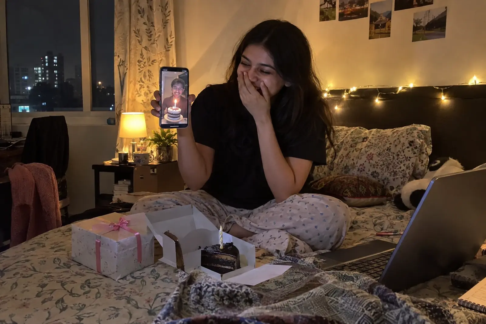 A young woman on her bed at midnight laughs at a video call where her boyfriend in another city holds up a cake