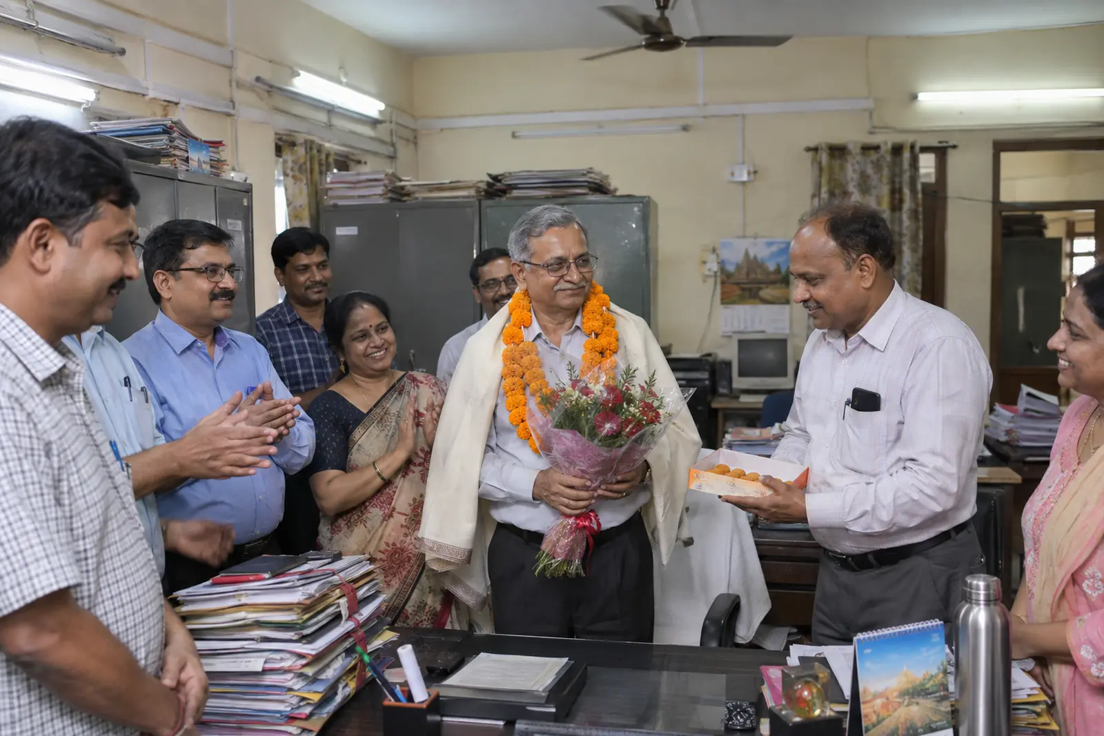 A man in a marigold garland and shawl holds a bouquet at his desk as colleagues offer sweets on his retirement day